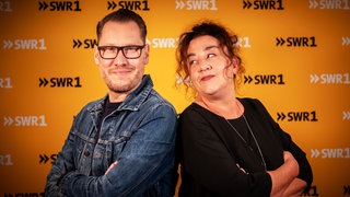 Die SWR1 Moderatoren Jochen Stöckle und Annett Lorisz stehen mit dem Rücken aneinander gelehnt und lächeln in die Kamera.