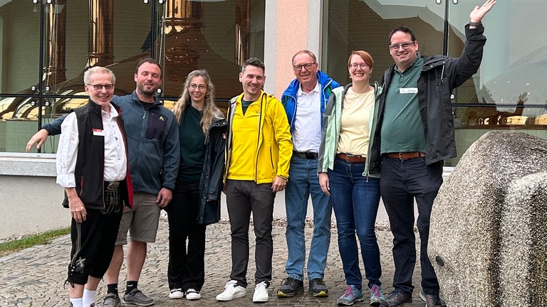 Gruppe von sieben Menschen steht vor einer Bierbrauerei und winkt
