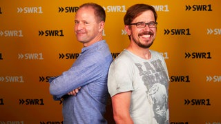 Die SWR1 Moderatoren Patrick Neelmeier und Ingo Lege stehen mit dem Rücken aneinandergelehnt und lächeln in die Kamera.
