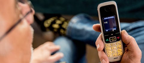 Enkeltrick oder falscher Polizist: Ein Mann sitzt in seiner Wohnung und hält ein Mobiltelefon in der Hand auf dem Display des Telefons ist die 110 zu lesen.