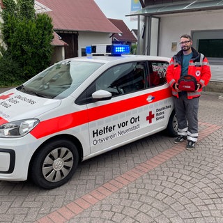 Daniel Müller steht neben dem Einsatzwagen von "Helfer vor Ort" - er hält einen Defibrillator in der Hand.