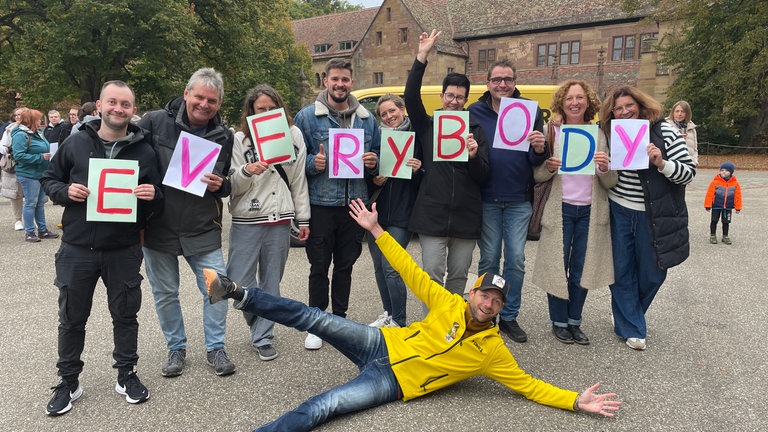 Gruppe hält schilder mit der Aufschrift Everybody in den Händen. Moderator Corvin Tondera-Klein liegt davor.