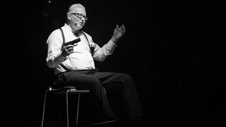 Schwarz-Weiss-Foto von Matthias Holtmann. Er sitzt bei Pop und Poesie 2012 auf der Bühne auf einem Stuhl. Dabei hält er eine Fake-Pistole in der Hand.