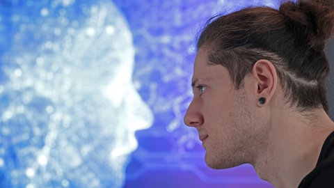 Ein Student sitzt im Computerraum der Hochschule vor einem Bildschirm mit einem schemenhaften Gesicht aus Lichtpunkten. Künstliche Intelligenz erkennt Krebs und Herzfehler und sortiert Kirschen. Ist sie auch gefährlich? So verändern Chat GPT und Co. unseren Alltag.