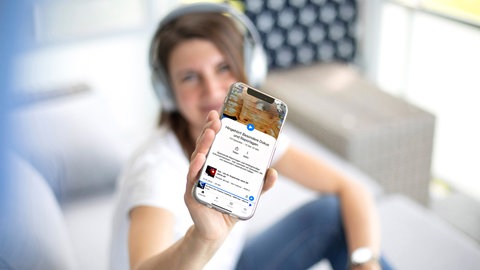 Frau im Hintergrund zeigt ein Smartphone mit der ARD Sounds App im Vordergrund. 