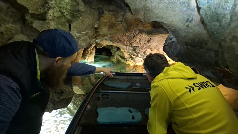 SWR1 Moderator Julian Hammerstein mit Bootsführer Valentin auf einem Boot in der Wimsener Höhle. Sie ziehen die Köpfe ein, da die Steine sehr tief kommen.