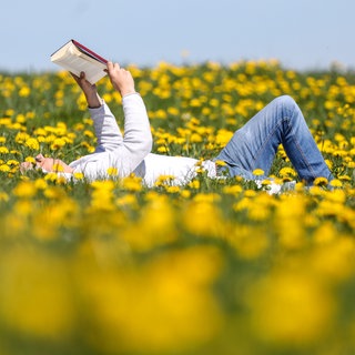Ein Mann liegt in einer Wiese mit blühendem Löwenzahn in der Sonne und liest ein Buch. Wir haben die besten Bücher und Buchtipps für euch.