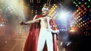 Freddie Mercury steht bei seinem Konzert in Mannheim 1986 auf der Bühne und trägt eine Krone und einen königlichen Umhang