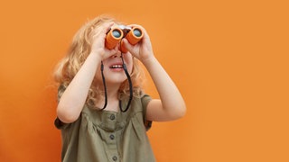 Ein Mädchen steht vor einer orangenen Wand und blickt mit einem orangenen Fernglas nach oben.