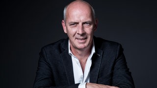 Ex-Fußballprofi Mario Basler steht mit verschränkten Armen vor einer Wand.