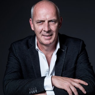 Ex-Fußballprofi Mario Basler steht mit verschränkten Armen vor einer Wand.