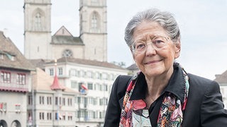 Gret Haller, ehemalige Diplomatin, blickt lächelnd in die Kamera. Im Hintergrund sind zwei Kirchtürme zu sehen.