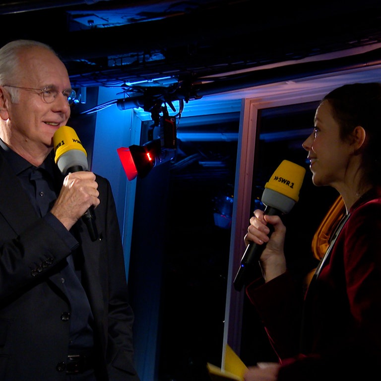 Entertainer Harald Schmidt und SWR1 Leute-Moderatorin Nicole Köster stehen zu gegenüber. Beide halten ein Mikrophon in der Hand und lächeln sich an.