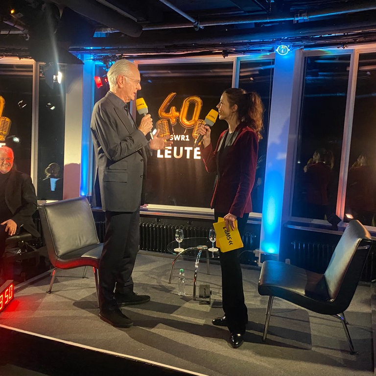 Auf einer kleinen Bühne stehen sich Entertainer Harald Schmidt und SWR1 Leute-Moderatorin Nicole Köster gegenüber. Beide halten ein SWR1 Mikrophon vor den Mund und sehen sich an. Im Hintergrund ist der Schriftzug zu 40 Jahre SWR1 Leute zu sehen.