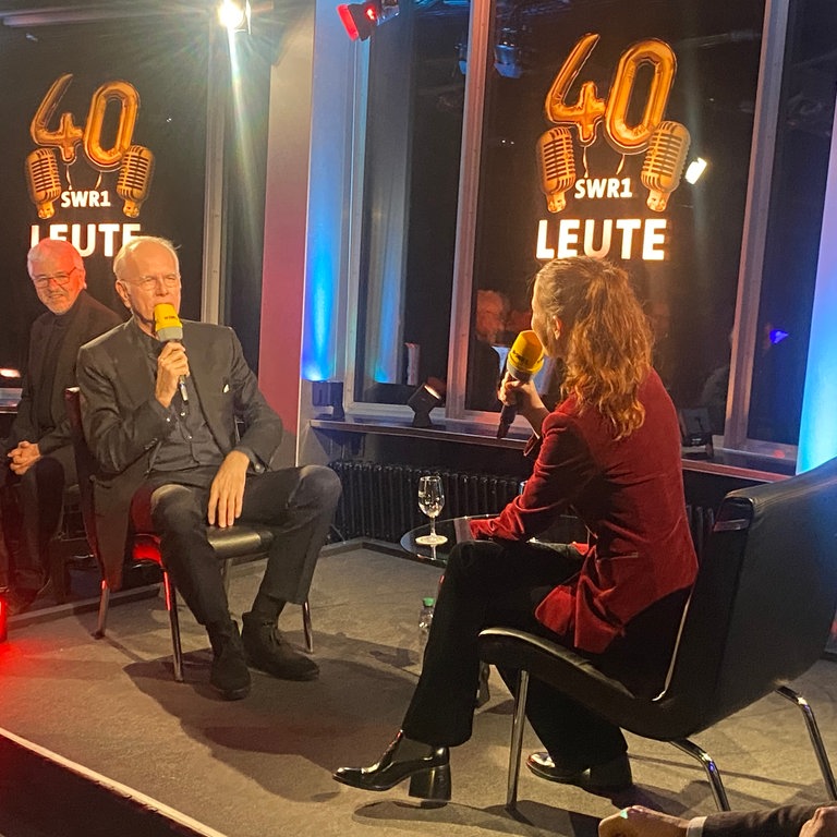 Entertainer Harald Schmidt und SWR1 Leute-Moderatorin Nicole Köster sitzen sich auf einer kleinen Bühne gegenüber. Beide haben ein Mikrophon vor dem Mund. Schmidt spricht.