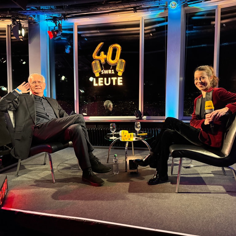 Entertainer Harald Schmidt und SWR1 Leute-Moderatorin Nicole Köster sitzen sich auf einer kleinen Bühne gegenüber. Beide sitzen zum Publikum gewandt und Schmidt hebt die linke Hand ans Ohr. Beide lächeln. Nicole Köster hält ein Mikrophon in der Hand.