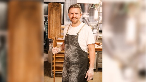 Ingmar Krimmer, Bäcker des Jahres 2024, über die Liebe zu Brot und Brezel. Er steht mit bemehlten Händen und Schürze in der Backstube und hält einen Brotschieber in der rechten Hand.