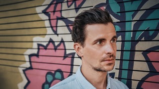 Julian Sengelmann, Schauspieler, Musiker, Theologe und neue SWR1 Station-Voice, steht vor einem Blumen-Graffiti.