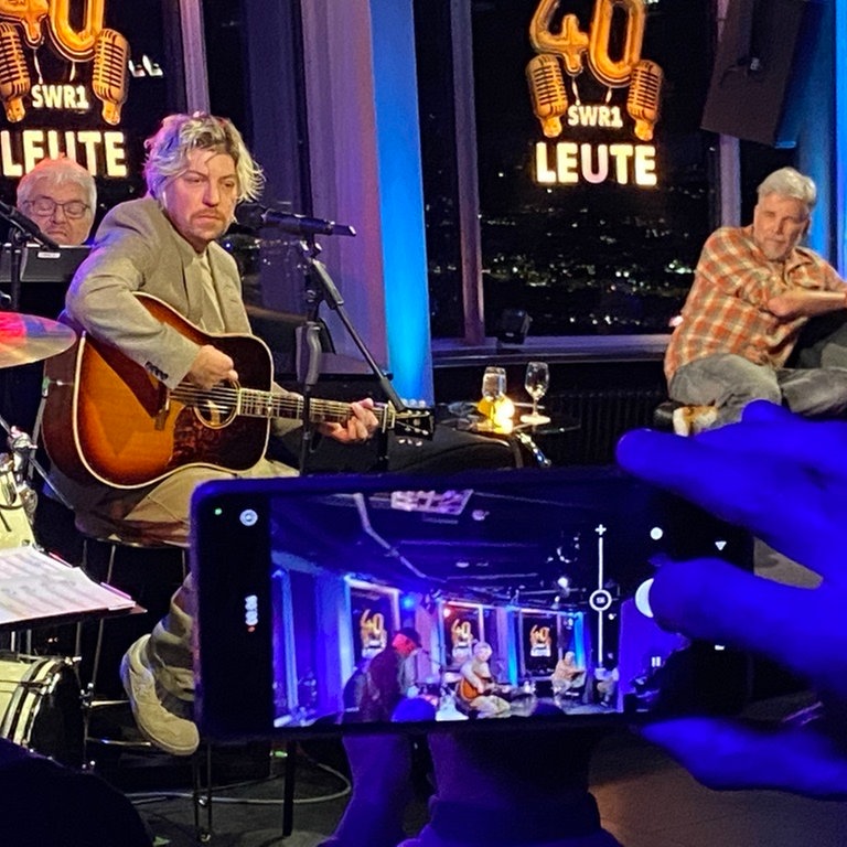 Sänger Philipp Poisel sitzt auf einem Hocker, spielt Gitarre und blickt ins Publikum. Am Rand schaut ihm SWR1 Leute-Moderator Jens Wolters zu. Im Vordergrund sieht man eine Hand mit Smartphone. Darauf abgebildet, die Szene auf der Bühne mit dem Sänger. 