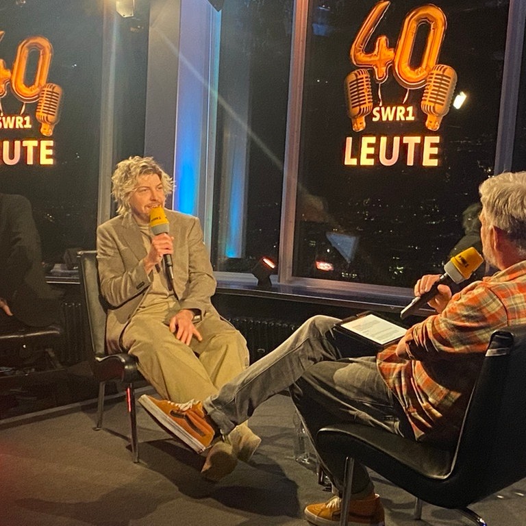 Sänger Philipp Poisel im Gespräch mit SWR1 Leute-Moderator Jens Wolters. Poisel sitzt hinten, spricht ins Mikrophon und sieht Wolters an. Dieser sitzt mit dem Rücken zum Betrachter, hat die Beine überschlagen und auf den Knien liegt ein Tablet. 