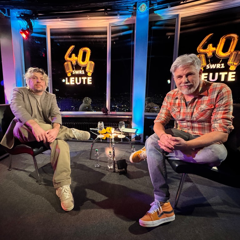 SWR1 Leute-Moderator Jens Wolters und Philipp Poisel sitzen auf einer kleinen Bühne. Sie sind dem Betrachter zugewandt und schauen freundlich entspannt. Im Hintergrund ist das Logo zu 40 Jahre SWR1 Leute. 