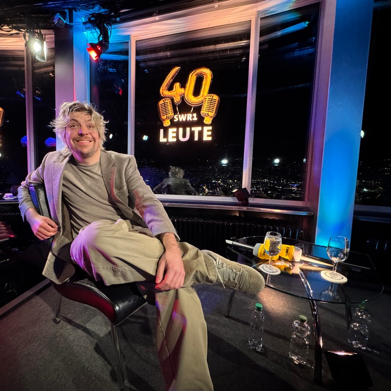 Sänger Philipp Poisel sitzt lässig auf der Bühne und lächelt dem Betrachter zu. Die Beine hat er überschlagen, eine Hand liegt entspannt auf dem Bein. Im Hintergrund ist das Logo zu 40 Jahre SWR1 Leute. 