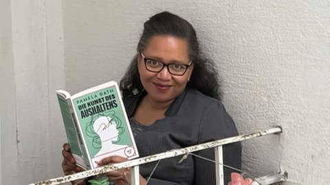 Philologin Pamela Rath sitzt auf einem Balkon und hält ihr Buch in der Hand.. In SWR1 Leute spricht sie darüber, wie uns Resilienz und Aushalten in Zeiten von Krisen und Kriegen helfen kann.