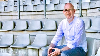 Alexander Jacob sitzt auf einem Tribünenplatz in einem Fußballstadion. Alexander Jacob litt jahrelang unter Wettsucht und klärt heute über Sportwetten und Spielsucht auf.