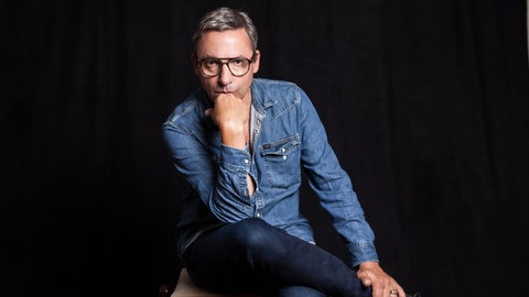 Musiker und Schriftsteller Rocko Schamoni sitzt mit übergeschlagenem Bein und auf die Hand aufgestütztem Kopf auf einem Stuhl. Die "Rocko Schamoni Supershow" trendet in der ARD Mediathek, seine "Dorfpunks" wurden zum Kultbuch, 20 Jahre später kommt mit "Pudels Kern" die Fortsetzung. Rocko Schamoni nimmt uns in SWR1 Leute mit in die 80er und 90er, zu den Toten Hosen, Bela B., Peter Maffay, Helge Schneider und in den legendären Hamburger Pudelclub.