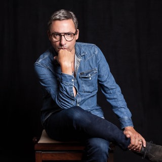 Musiker und Schriftsteller Rocko Schamoni sitzt mit übergeschlagenem Bein und auf die Hand aufgestütztem Kopf auf einem Stuhl. Die "Rocko Schamoni Supershow" trendet in der ARD Mediathek, seine "Dorfpunks" wurden zum Kultbuch, 20 Jahre später kommt mit "Pudels Kern" die Fortsetzung. Rocko Schamoni nimmt uns in SWR1 Leute mit in die 80er und 90er, zu den Toten Hosen, Bela B., Peter Maffay, Helge Schneider und in den legendären Hamburger Pudelclub.