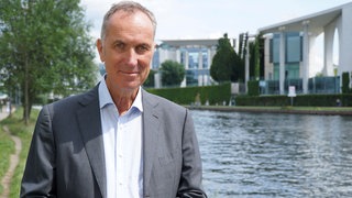 Stephan Lamby steht an einem Kanal, dahinter sind verschwommen Häuser zu erkennen.