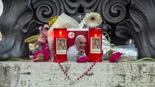 Ein Bild des verstorbenene Papstes Franziskus steht inmitten von Kerzen und Blumen. Ein Rosenkranz ist um das Bild geschlungen. Trauerfeierlichkeiten, Beisetzung, Papst-Wahl im Konklave – ein minutiös durchgeplantes Räderwerk ist nach dem Tod von Papst Franziskus im Vatikan in Bewegung. Vatikan-Experte Andreas Englisch erklärt es in SWR1 Leute im Detail.