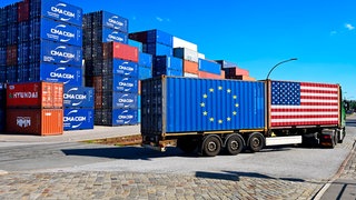 Container mit Europa- und USA-Fahne auf einem LKW. Im Hintergrund stapeln sich Container. Symbolbild für Zollstreit und Zoll-Deal zwischen EU und USA.
