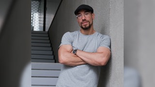 Maximilian Pollux Autor, Podcaster, Anti-Gewalttäter und ehemaliger Gangster, lehnt mit verschränkten Armen an einer Wand.