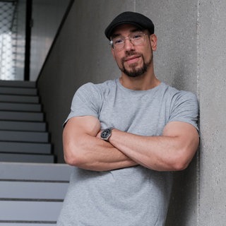 Maximilian Pollux Autor, Podcaster, Anti-Gewalttäter und ehemaliger Gangster, lehnt mit verschränkten Armen an einer Wand.