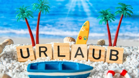 Das Wort "Urlaub" steht aus Holzbuchstaben im Sand, umgeben von Palmen, Muscheln und einem kleinen Boot. In SWR1 Leute spricht Reisereporter Christoph Karrasch über Reisetrends und Urlaubsländer 2026.