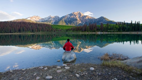 Ein Mann sitzt in einem See auf einem großen Stein und blickt auf ein Bergpanorama. In SWR1 Leute spricht Wolfgang Fasser über die große Bedeutung von Ruhe und Stille für uns.