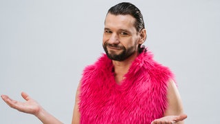 Martin Puntigam, Kabarettist und Schauspieler, dreht in pinker Puschelweste die Handflächen nach oben. Er kombiniert Topwissenschaft und Spitzenhumor.