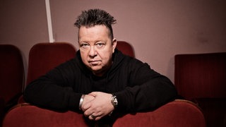 Musiker Sebastian Krumbiegel lehnt mit den Armen auf den Rückenlehnen der vorderen Sitzreihe.