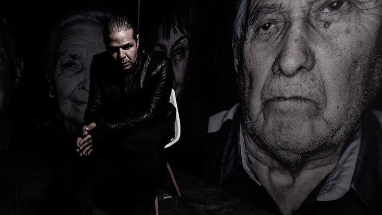 Fotograf Luigi Toscano sitzt auf einem Stuhl, dahinter drei Portaits von Holocaust-Überlebenden.