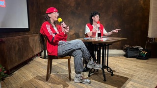 An einem Tisch sitzen Yvonne und Ria mit roten Jacken über weißen T-Shirts. Sie halten Mikrofone in den Händen.