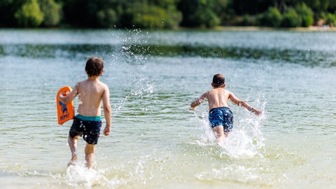 Zwei Kinder rennen im Sommer an einem Badesee bei Berum vom Ufer ins Wasser. Sommer, Hitze und der Wunsch nach Abkühlung: In diesen Freibädern, Baggerseen und Flüssen in Baden-Württemberg ist Baden und Schwimmen erlaubt.