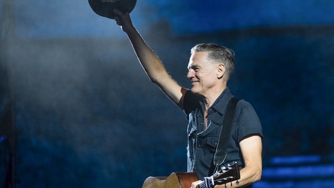 Bryan Adams hebt bei einem Akustikkonzert in Pompeji in Italien lächelnd seinen Hut an. Die Gitarre hält er mit der anderen Hand fest. (Juli 2025) | "Roll With The Punches" – das neue Album von Bryan Adams