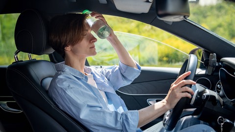 Eine Frau sitzt im Auto und hält sich eine Wasserflasche gegen die Stirn | Gesundheitsrisiko im Sommer: Das könnt ihr bei Hitze im Auto tun