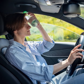 Eine Frau sitzt im Auto und hält sich eine Wasserflasche gegen die Stirn | Gesundheitsrisiko im Sommer: Das könnt ihr bei Hitze im Auto tun