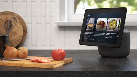 Schicker Smart Speaker mit Bildschirm und Alexa zu gewinnen: Ein Anthrazit-farbener Smart Speaker mit Display steht auf einer Arbeitsplatte in der Küche vor dem Fenster. Daneben liegen Zwiebeln und Tomaten auf einem Schneidebrett.