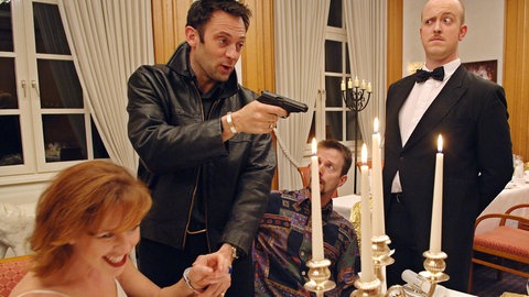 Krimidinner zuhause macht Spaß: Mehrere Personen sitzen oder stehen um einen feierlich gedeckten Tisch und spielen ihre Rollen. Einer hält eine Pistole in der Hand, eine Frau trägt Handschellen, der nächste guckt verduzt. 