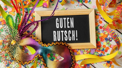 Silvesterparty: Auf einem Tisch sieht man Konfetti und Luftschlangen und eine glitzernde Maske. Dazischen liegt eine kleine Tafel, auf der geschrieben steht: Guten Rutsch.