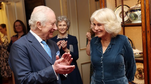 Links steht König Charles im Anzug und klatscht für Königin Camilla, die rechts im dunkelblauen Kleid steht. Beide strahlen übers ganze Gesicht.