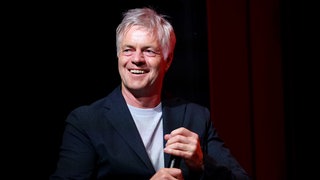 Sportkommentator Tom Bartels sitzt auf einem roten Stuhl mit einem Mikro in der Hand und lacht.